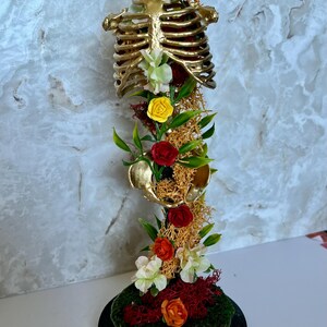 May include: A gold-colored, decorative rib cage sculpture adorned with colorful artificial flowers and greenery. The sculpture is mounted on a black, round base and features red, yellow, and white flowers, with green leaves and moss.