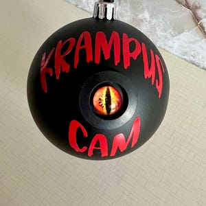 May include: A matte black spherical ornament featuring the red text KRAMPUS CAM and a central inset glass eye with a yellow iris and a vertical black pupil. The ornament has a silver metal cap and a black ribbon loop for hanging purposes.