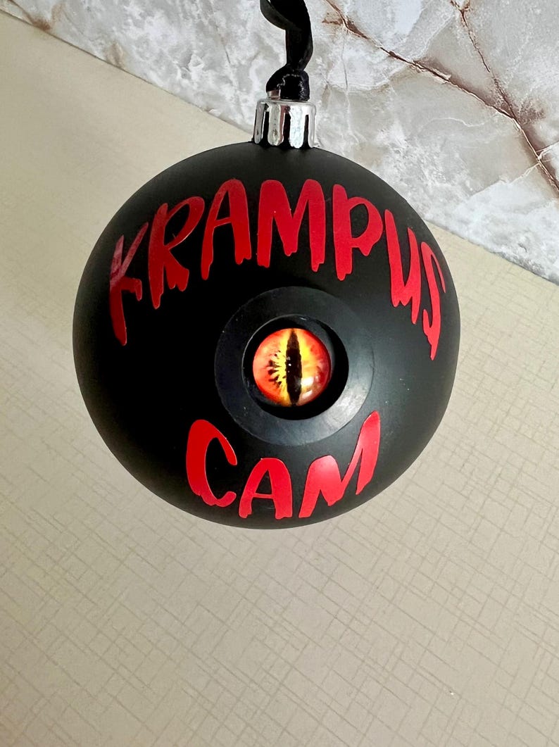 May include: A matte black spherical ornament featuring the red text KRAMPUS CAM and a central inset glass eye with a yellow iris and a vertical black pupil. The ornament has a silver metal cap and a black ribbon loop for hanging purposes.