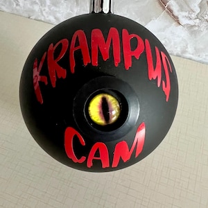 May include: A matte black spherical ornament featuring the red text KRAMPUS CAM printed on the front. Centered between the words is a realistic, glossy yellow and black reptilian eye set within a circular black frame, topped with a silver cap.