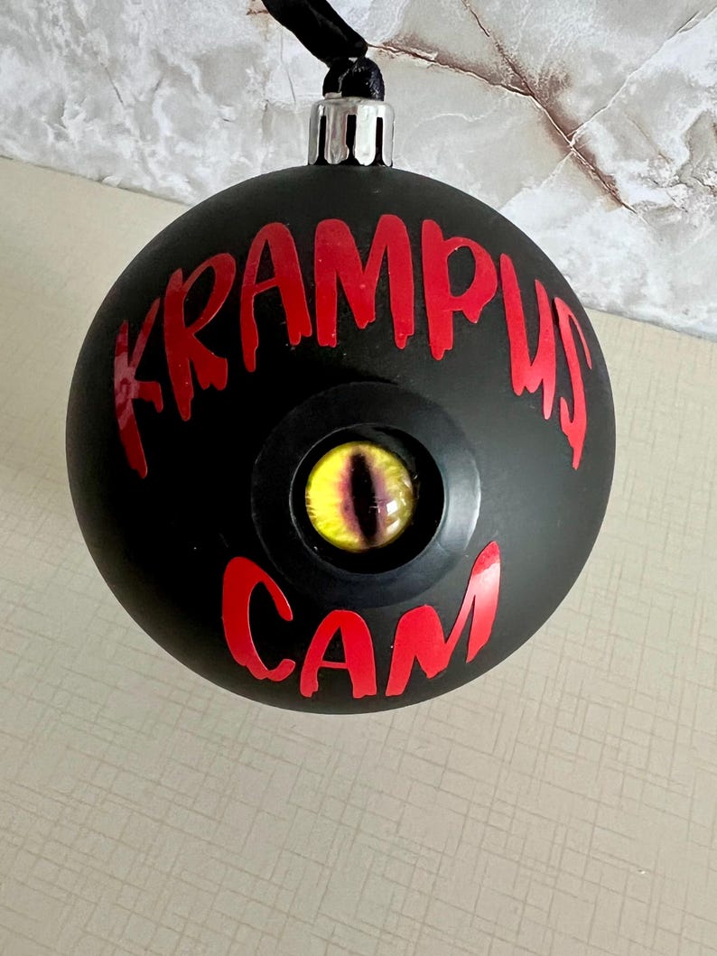 May include: A matte black spherical ornament featuring the red text KRAMPUS CAM printed on the front. Centered between the words is a realistic, glossy yellow and black reptilian eye set within a circular black frame, topped with a silver cap.