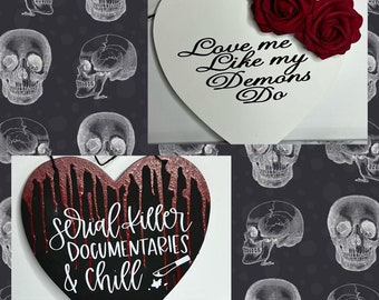 May include: Two black and red heart-shaped signs with white lettering. The first sign says "Love me like my demons do". The second sign says "Serial killer documentaries & chill" with a small knife graphic.