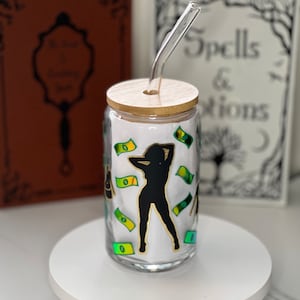 May include: A clear glass tumbler with a bamboo lid and a clear glass straw. The tumbler features a black silhouette of a person with gold accents, surrounded by green and yellow dollar bill graphics. The background includes two books.
