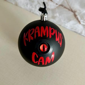 May include: A matte black spherical ornament featuring the red text KRAMPUS CAM and a central, realistic red and orange eye with a vertical black pupil. The ornament has a silver metal cap at the top with a black hanging loop attached.