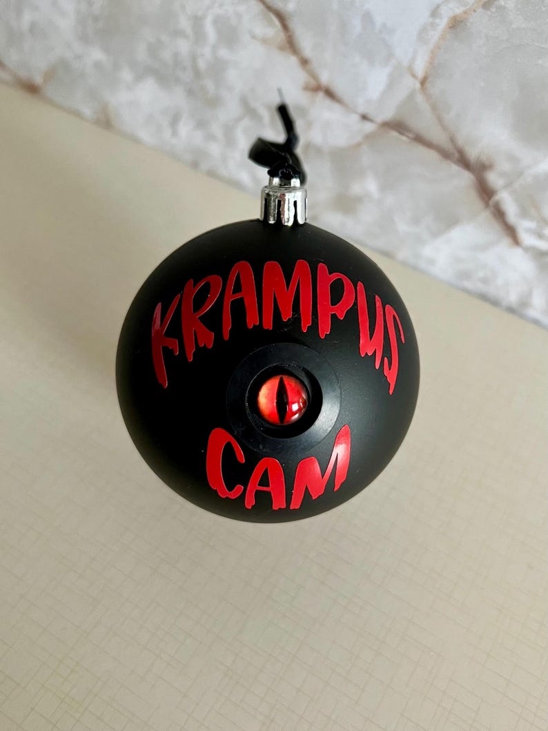 May include: A matte black spherical ornament featuring the red text KRAMPUS CAM and a central, realistic red and orange eye with a vertical black pupil. The ornament has a silver metal cap at the top with a black hanging loop attached.