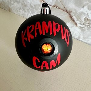 May include: A matte black spherical Christmas ornament featuring the red text KRAMPUS CAM printed on the front. Centered between the words is a circular inset containing a realistic, glowing orange and yellow reptilian eye with a vertical pupil.