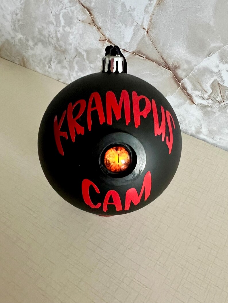 May include: A matte black spherical Christmas ornament featuring the red text KRAMPUS CAM printed on the front. Centered between the words is a circular inset containing a realistic, glowing orange and yellow reptilian eye with a vertical pupil.