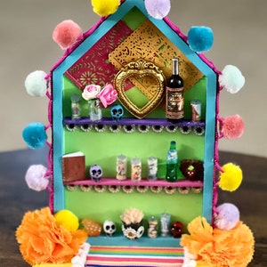 May include: A miniature wooden shelf with a green background, decorated with colorful pom-poms, paper flowers, and a heart-shaped frame. The shelf is filled with miniature items, including a bottle of wine, candles, and skulls. The shelf is sitting on a yellow base with a striped rug.
