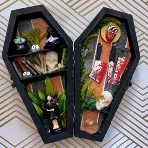 May include: A black coffin-shaped display box, open to reveal two compartments filled with miniature Halloween-themed items. Includes a skull, fairy figurine, miniature books, cleavers, and a witch hat. The background has a gold and white geometric pattern.