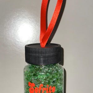 May include: A small glass jar filled with green and clear glass beads. The jar has a black lid and a red ribbon attached to the lid. The jar has the word "Sofrito" printed on the front in red.