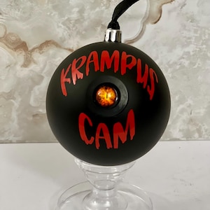 May include: A matte black spherical ornament featuring the red text KRAMPUS CAM and a central, realistic orange and black reptilian eye. The ornament has a silver cap and a black ribbon loop for hanging, displayed on a clear pedestal stand.