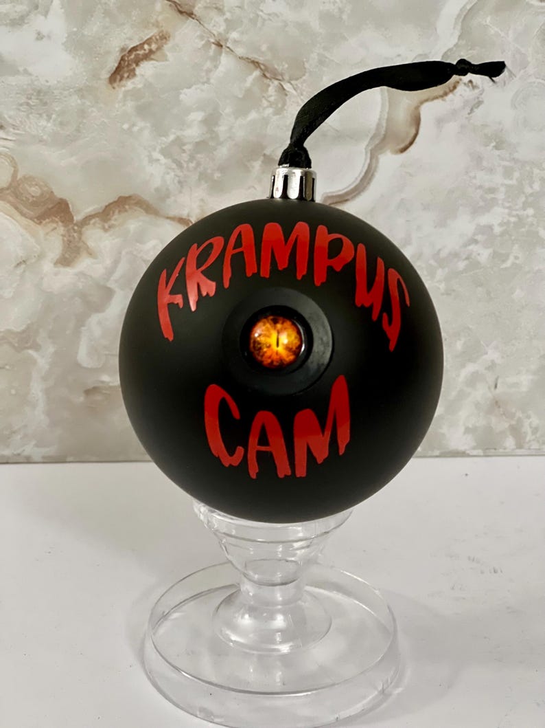 May include: A matte black spherical ornament featuring the red text KRAMPUS CAM and a central, realistic orange and black reptilian eye. The ornament has a silver cap and a black ribbon loop for hanging, displayed on a clear pedestal stand.