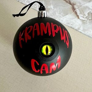 May include: A matte black spherical ornament featuring the red text KRAMPUS CAM and a central, realistic yellow eye with a vertical black pupil. The ornament includes a silver cap and a black hanging string for display purposes.