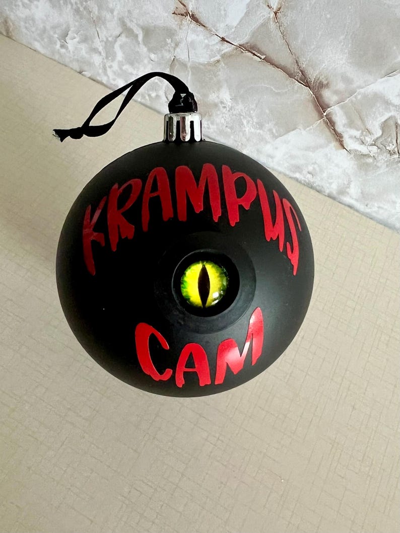 May include: A matte black spherical ornament featuring the red text KRAMPUS CAM and a central, realistic yellow eye with a vertical black pupil. The ornament includes a silver cap and a black hanging string for display purposes.