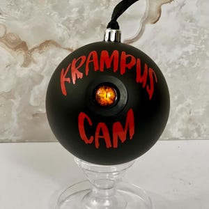 May include: A matte black spherical ornament featuring the red text KRAMPUS CAM and a central, realistic orange and black eye graphic. The ornament has a silver metal cap and a black ribbon loop for hanging, designed with a spooky aesthetic.