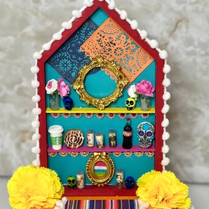 May include: A colorful Day of the Dead altar with a turquoise background, red trim, and white pom-poms. The altar has three shelves with various decorations, including a gold frame, flowers, candles, and a Coca-Cola bottle. The altar is decorated with yellow marigolds and a colorful striped rug.