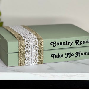 May include: Two stacked, sage green, decorative boxes with a burlap and lace ribbon. The top box says "Country Roads" and the bottom box says "Take Me Home".