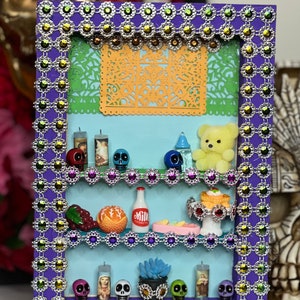 May include: A decorative shadow box with a purple frame and shelves, adorned with sparkling silver and jewel accents. The shelves display miniature Day of the Dead skulls, candles, a teddy bear, and other colorful figurines. A cut-out orange paper design is in the background.