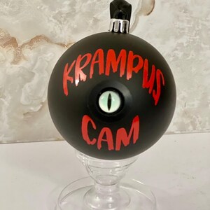 May include: A matte black spherical ornament featuring the red text KRAMPUS CAM and a central, realistic green reptilian eye with a vertical black pupil. The ornament has a silver metal cap at the top and rests on a clear plastic stand.