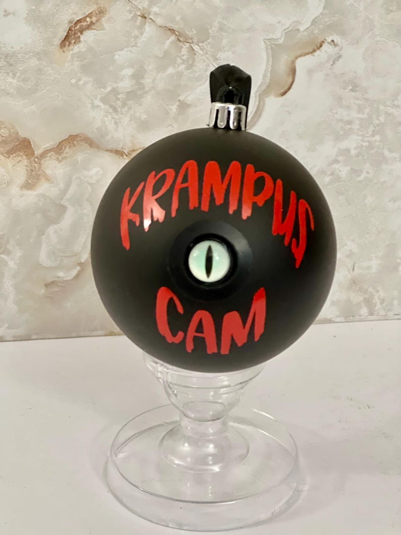 May include: A matte black spherical ornament featuring the red text KRAMPUS CAM and a central, realistic green reptilian eye with a vertical black pupil. The ornament has a silver metal cap at the top and rests on a clear plastic stand.