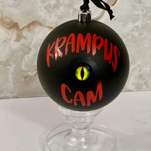 May include: A matte black spherical ornament featuring the red text KRAMPUS CAM and a central, realistic yellow and black reptilian eye. The ornament has a silver metal cap with a black ribbon loop and rests on a clear, pedestal-style stand.