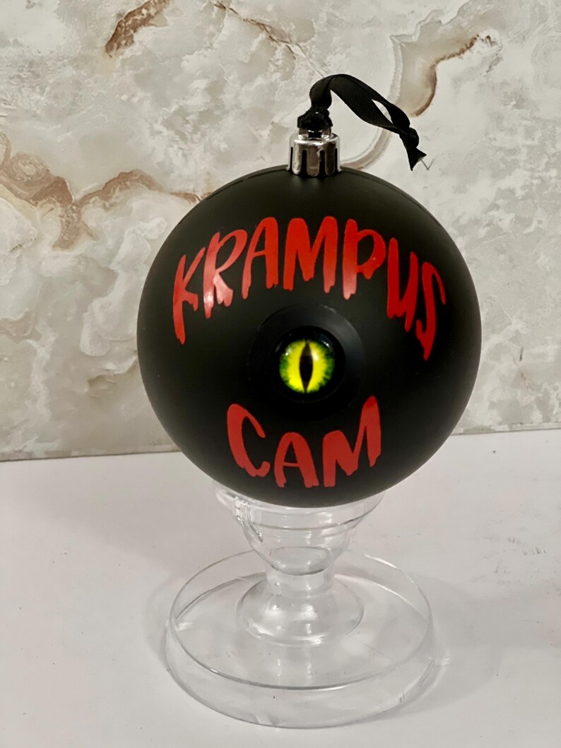 May include: A matte black spherical ornament featuring the red text KRAMPUS CAM and a central, realistic yellow and black reptilian eye. The ornament has a silver metal cap with a black ribbon loop and rests on a clear, pedestal-style stand.
