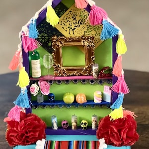 May include: A miniature Day of the Dead altar with a green and purple wooden frame, decorated with colorful paper flowers, tassels, and skulls. The altar has two shelves with various miniature items, including a bottle of wine, a glass, a framed picture, and candles.