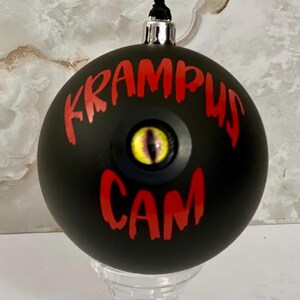 May include: A matte black spherical ornament featuring the red text KRAMPUS CAM and a central yellow reptilian eye with a vertical black pupil. The ornament has a silver metal cap and a black ribbon loop for hanging from a tree branch.