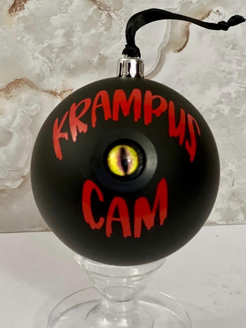 May include: A matte black spherical ornament featuring the red text KRAMPUS CAM and a central yellow reptilian eye with a vertical black pupil. The ornament has a silver metal cap and a black ribbon loop for hanging from a tree branch.