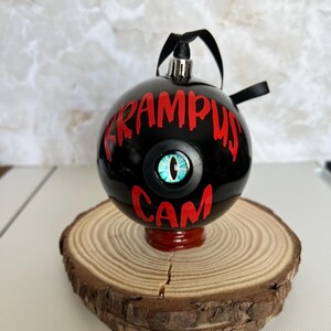 May include: A spherical black Christmas ornament featuring the red text KRAMPUS CAM and a central blue reptilian eye graphic. The ornament has a silver cap with a black ribbon loop for hanging, designed with a glossy finish and bold lettering.