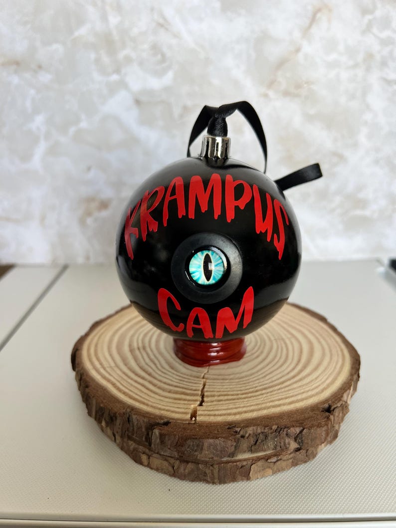 May include: A spherical black Christmas ornament featuring the red text KRAMPUS CAM and a central blue reptilian eye graphic. The ornament has a silver cap with a black ribbon loop for hanging, designed with a glossy finish and bold lettering.