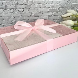 May include: A pink gift box with a clear lid and a pink satin ribbon. The box is tied with a bow and has a marbled pattern on the inside.