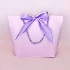 May include: A lavender tote bag with a matching satin ribbon bow and rope handles. The bag is a solid color and has a simple, elegant design. The background is a soft pink.