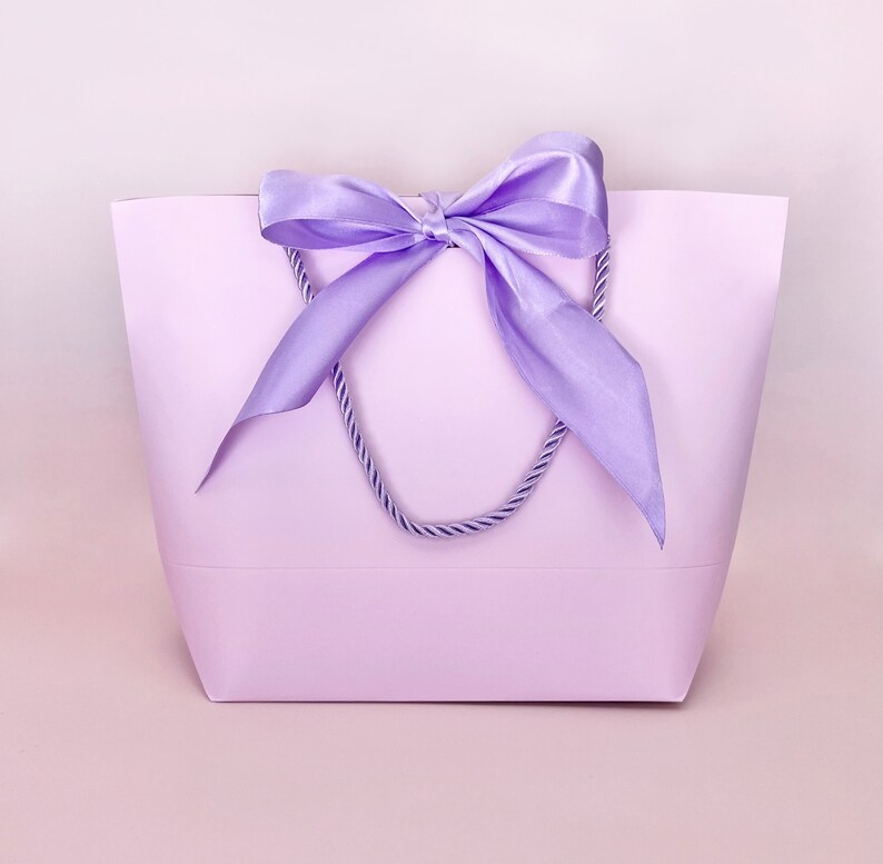 May include: A lavender tote bag with a matching satin ribbon bow and rope handles. The bag is a solid color and has a simple, elegant design. The background is a soft pink.