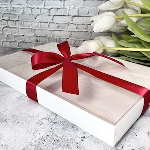 May include: A white gift box with a clear lid, tied with a red satin ribbon and bow. The box is sitting on a gray textured surface.