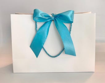 May include: A white gift bag with a bright blue satin bow and matching rope handles. The bow is centered on the front of the bag, adding a decorative touch. The bag is clean and simple, suitable for various occasions.