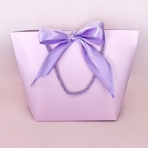 May include: A lavender gift bag with a matching satin bow and rope handles. The bag is a solid color, with the bow tied at the top center. The background is a soft pink.