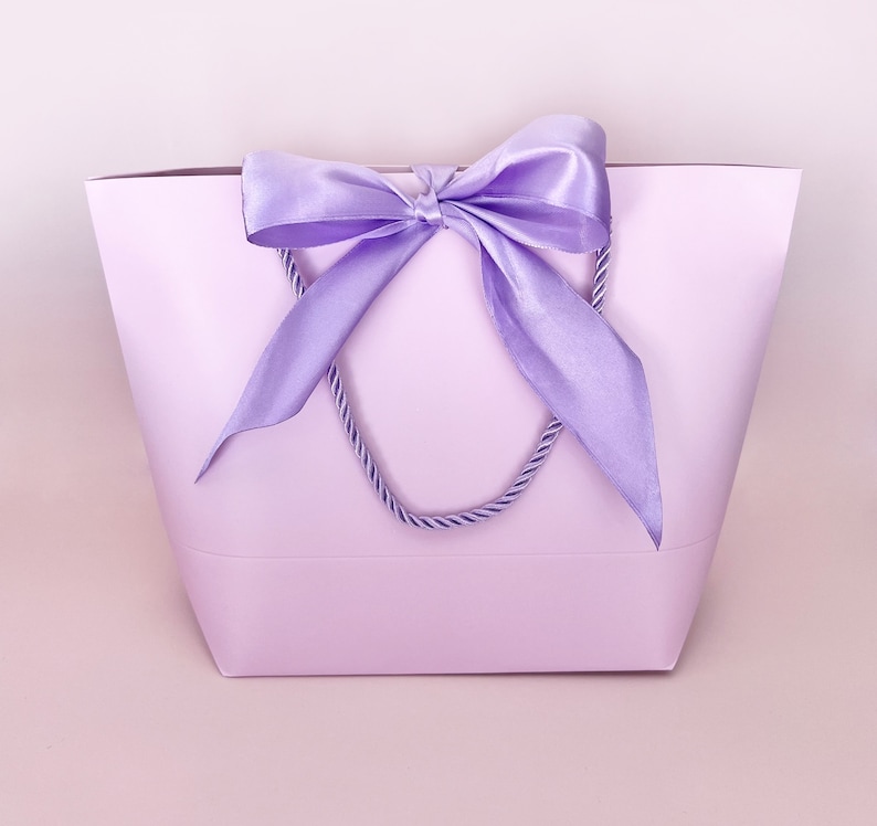 May include: A lavender gift bag with a matching satin bow and rope handles. The bag is a solid color, with the bow tied at the top center. The background is a soft pink.