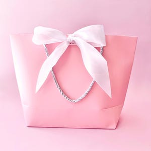 May include: A pink gift bag with a white satin bow and silver rope handles. The bag is a solid pastel pink color, and the bow is tied at the top center. The background is a matching pink.