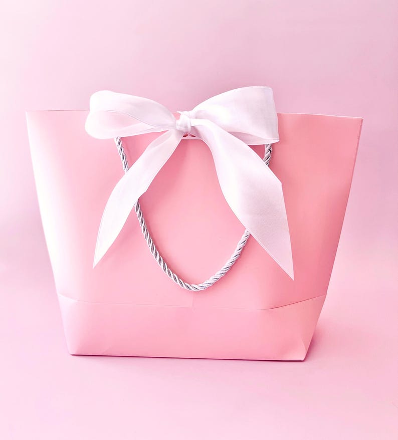 May include: A pink gift bag with a white satin bow and silver rope handles. The bag is a solid pastel pink color, and the bow is tied at the top center. The background is a matching pink.