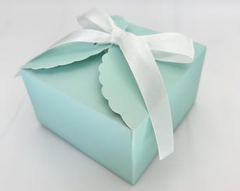 May include: A small, light blue gift box with a white satin ribbon tied in a bow. The box has a scalloped edge on the lid and is closed with the ribbon. The box is sitting on a white surface.