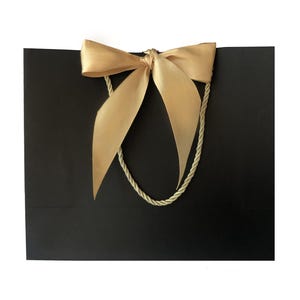 May include: A black gift bag with a gold satin bow and matching rope handles. The bag is rectangular and has a simple, elegant design. The bow is tied at the top center of the bag.