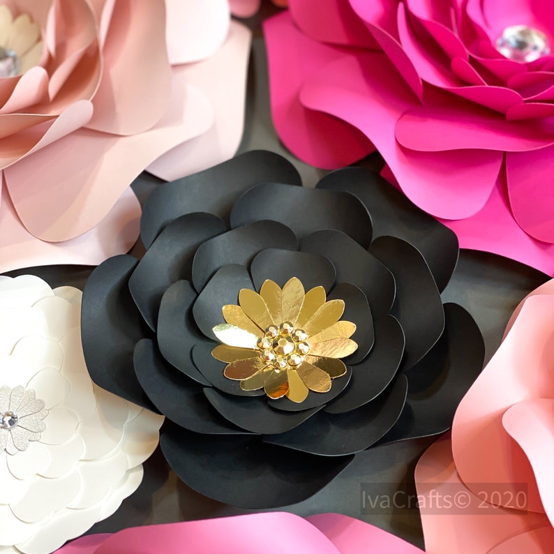 May include: A close-up of three large paper flowers. The flowers are black, pink, and white. The black flower has a gold center with a rhinestone embellishment. The pink flower has a rhinestone embellishment. The white flower has a silver rhinestone embellishment. The flowers are arranged in a cluster.