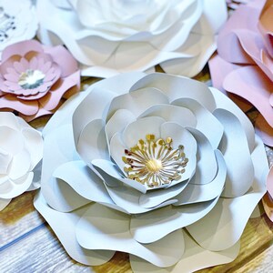 May include: A large, light blue paper flower with a gold center. The flower is made of multiple layers of paper and has a delicate, ruffled texture. The flower is surrounded by other paper flowers in shades of pink and white.