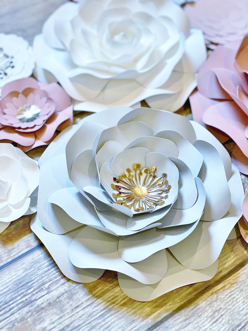 May include: A large, light blue paper flower with a gold center. The flower is made of multiple layers of paper and has a delicate, ruffled texture. The flower is surrounded by other paper flowers in shades of pink and white.
