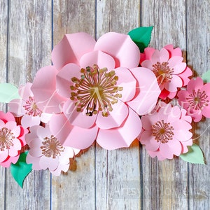 May include: A set of pink paper flowers with gold centers. The flowers are arranged in a cluster on a wooden background.