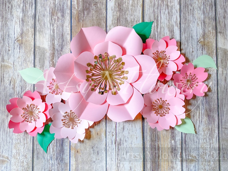 May include: A set of pink paper flowers with gold centers. The flowers are arranged in a cluster on a wooden background.