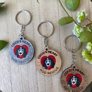 Puede incluir: Tres llaveros de madera con una silueta de perro dentro de un corazón rojo. Cada llavero tiene el texto "Coco's Heart Dog Rescue". Los llaveros están sobre una superficie de madera.