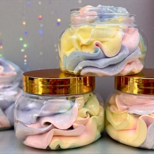 May include: Four clear jars filled with colorful, whipped soap. The soap is swirled with pastel shades of pink, yellow, blue, and purple. The jars have gold lids and are arranged on a light surface.