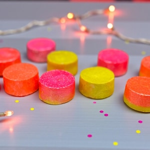 May include: A selection of brightly coloured, round, textured objects in pink, orange, and yellow. They are arranged on a light grey surface, with fairy lights and confetti adding a festive touch. The objects appear to be approximately 2.5 cm in diameter.